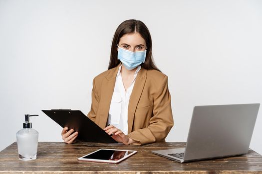 Covid, pandemic and office workplace concept. Portrait of corporate woman in medical face mask from coronavirus, working, standing over white background.