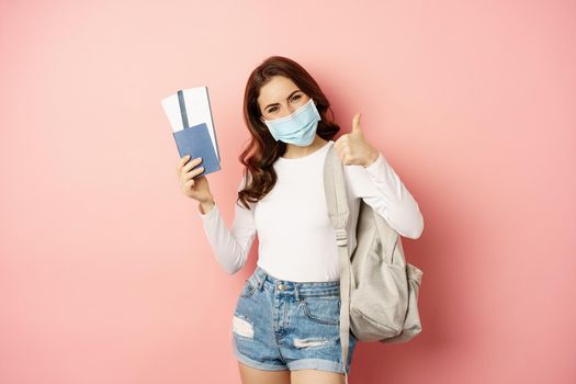 Covid and travelling concept. Happy woman traveller in medical mask, holding passport and airplane tickets, going on trip, vacation during quarantine, pink background.