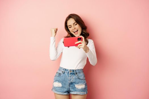 Beautiful girl looking enthusiastic, scream of joy and excitement, winning on mobile phone video game, watching smth on smartphone and celebrating, pink background.