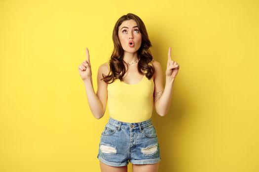 Trendy brunette girl looking and pointing fingers up, showing something upwards, checking out promo offer, store, deal, standing over yellow background.