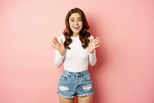 Image of excited smiling stylish woman, shaking hands and jumping enthusiastic, looking amazed at camera, standing against pink background.