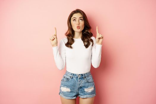 Portrait of surprised caucasian woman, stylish girl pointing fingers up and looking impressed, showing store logo, discount advertisement, pink background.