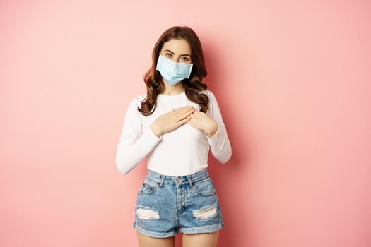 Portrait of beautiful girl in medical face mask, holding hands on heart and smiling, looking with appreciation and care, standing over pink background.