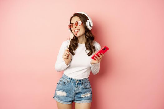 Stylish summer girl dancing with smartphone and lolipop, listening music in headphones, standing in sunglasses against pink background.