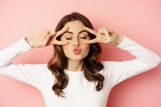 Close up face of young stylish girl, sassy woman showing happy, excited face expression, peace v-sign over eyes, standing against pink background.