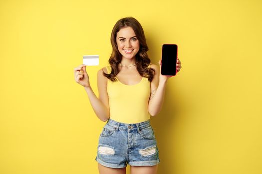 Young beautiful woman model showing smartphone empty phone screen and credit card, ready for summer, wearing tank top and shorts, standing over yellow background.