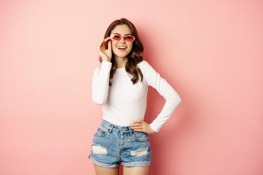 Portrait of stylish glam girl in sunglasses, laughing and smiling, standing over holiday pink background. Copy space