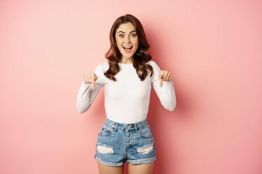 Enthusiastic beautiful and stylish girl showing advertisement, pointing fingers down at announcement and smiling, pink background.