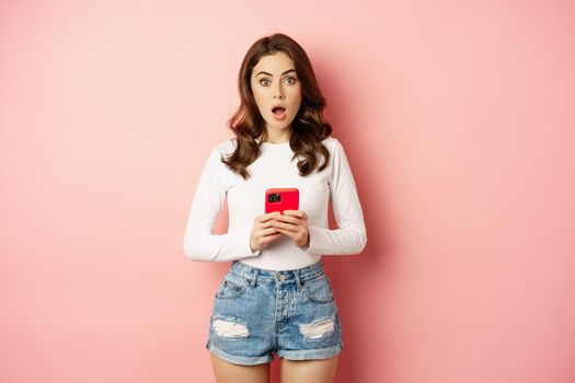 Surprised woman holding pink smartphone and gasping amazed, looking impressed and shocked at camera, studio background.