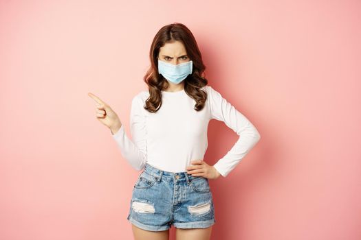 Angry young woman in medical face mask, frowning frustrated, pointing finger left, showing promo copy space, standing over pink background.