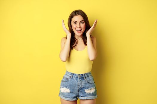 Portrait of happy girl open eyes, looking amazed at surprise and smiling, gift reveal, standing in summer outfit over yellow background.