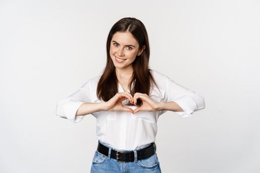 Lovely feminine woman showing heart sign, romantic gesture and smiling with care and tenderness, standing over white background.