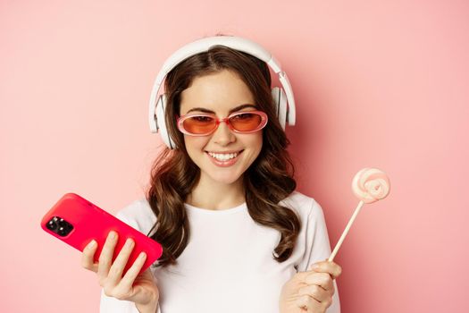 Close up portrait of stylish glamour girl, beautiful woman in headphones, listening music with smartphone, wearing sunglasses and licking lolipop.