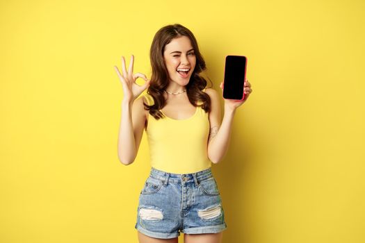 Pretty brunette woman, showing mobile phone screen, smartphone app interface and okay sign, recommend website, store, standing against yellow background.