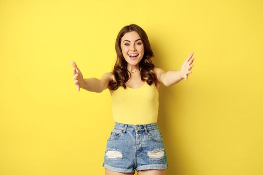 Beautiful girl in tank top, reaching hands, stretching arms forward and smiling, welcome, hugging or greeting someone, standing over yellow background.