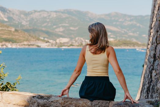 Beautiful girl sitting on a stone wall, in background is the blue sea, Budva, Montenegro