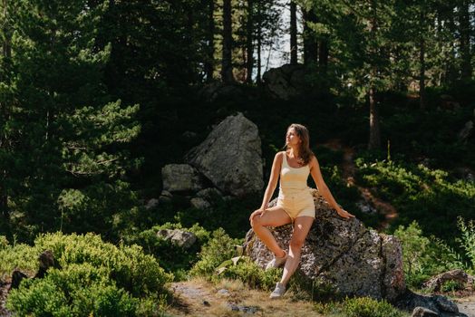 Young woman tourist sitting on the mountain peak and beautiful mountains. Landscape with sporty girl, green forest, hills , sky, lake. Travel and tourism. Hipster girl in the mountains. Stylish woman in checkered shirt sitting on stones in forest. Wanderlust concept. Hiking and travelling in summer. mysterious wood. Beautiful nature