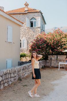 Girl tourist walking through ancient narrow street on a beautiful summer day in MEDITERRANEAN MEDIEVAL CITY , OLD TOWN bUDVA, MONTENEGRO. Young beautiful cheerful woman walking on old street at tropical town. Pretty girl looking at you and smiling