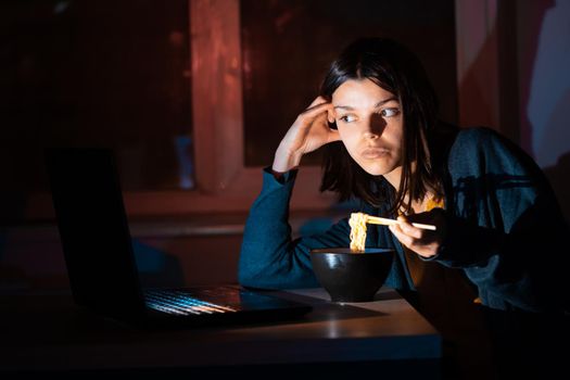 Young girl works at night at home with laptop and eats noodles, asian food with chopsticks. A woman watches a show, a movie and has dinner with ordered food from a cafe.