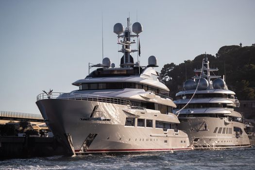 Two huge yachts are in port of Monaco at sunset, sun reflection are on glossy boards of motor boats, the chrome plated handrail, megayacht is moored in marina, mountain is on background. High quality photo