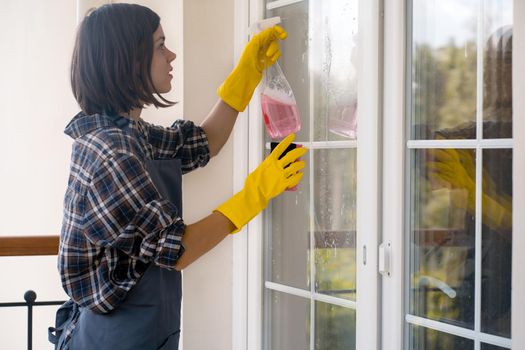 A young girl in an apron does housework, washes the windows with a special product, wipes it dry with a rag, a woman washes the glass with a sponge.