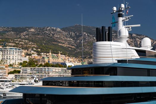 Landmarks of Monaco through decks of huge yacht of blue color in port Hercule at sunny day, glossy board of the motor boat, sun reflection on glossy board, mountains are on background. High quality photo