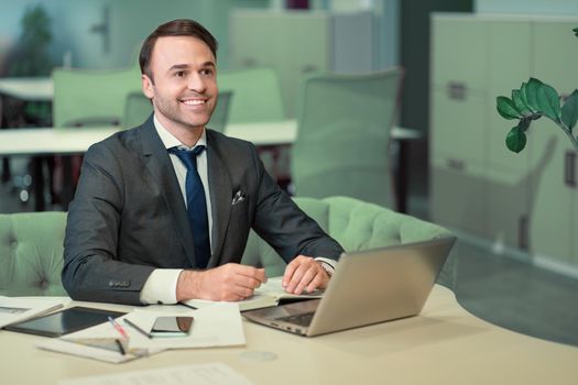 Handsome man in business suit working on laptop, job in progress. Cheer smiling freelancer making notes in scheduler sitting in bright coworking space.