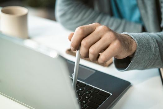 Close-up freelancer man working at laptop sitting at the desk at home typing using a pen. Remote working pensive young man working at home. Work from home concept.