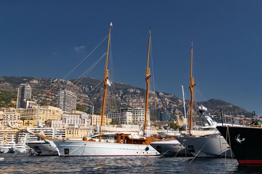 A lot of huge yachts are in port of Monaco at sunny day, Monte Carlo, mountain is on background, big masts of sail yacht, motorboat and mega yachts are moored in marina, sun reflection on glossy board. High quality photo