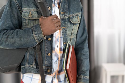African American man holding books and backpack strap wearing denim jacket with study books. Handsome young student man on the way to school. School concept. Studies concept. No face visible.