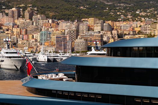 Landmarks of Monaco through decks of huge yacht of blue color in port Hercule at sunny day, glossy board of the motor boat, sun reflection on glossy board, mountains are on background. High quality photo