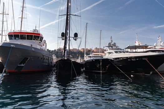 A lot of huge yachts are in port of Monaco at sunny day, Monte Carlo, mountain is on background, big masts of sail yacht, motorboat and mega yachts are moored in marina, sun reflection on glossy board. High quality photo