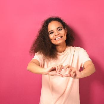 Sending love gesture beautiful young African American woman looking positively at camera wearing peachy t-shirt isolated on pink background. Beauty concept. Square crop.