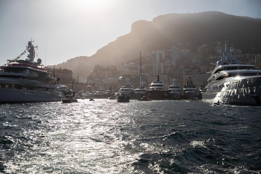 A lot of huge yachts are in port Hercule of Monaco at sunset, mountain is on background, magic backlight is on glossy boards of the motor boats, megayachts are moored in marina, sun reflections. High quality photo