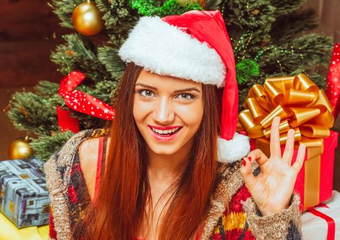 New Year Woman with Long Hair in a Red Hat Gazes to the Camera and Shows Hand Ok Sign. Green Decorated New Year Tree with lots of Gift Boxes Background. Close-up. High quality photo
