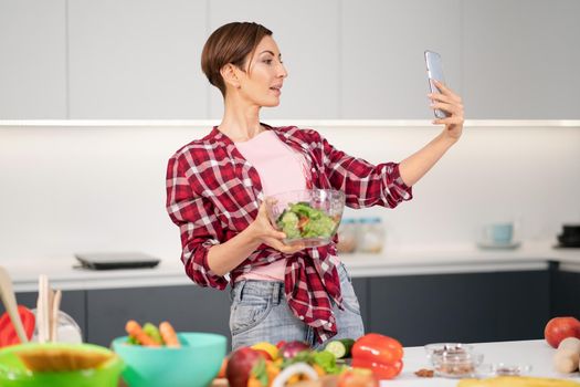 Pretty housewife taking selfie or making a video call using her smartphone while cooking fresh salad wearing a plaid shirt with a bob hair style. Healthy food leaving - vegan concept.