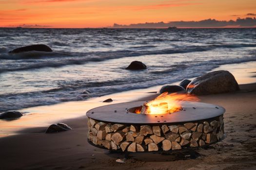 The beautiful metal round a fireplace stands on the sandy coast with a tidal wave at sunset, stones, sand, waves, the landscape, tranquillity, the sun falls, firewood, nobody. High quality photo