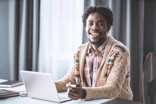 African man working from home using laptop sitting next to the window. Young entrepreneur working on his laptop at home. African american student study at home during lockdown.