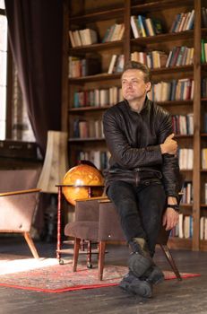 Brutal Guy of 35s in Biker Style Clothes Sits on the Arm of a Vintage Chair in the Middle of an Old Retro Room. Retro Interior, Bookcase, Floor Lamp, Antique world globe, Rustic Rug on the Floor. Close-up. High quality photo