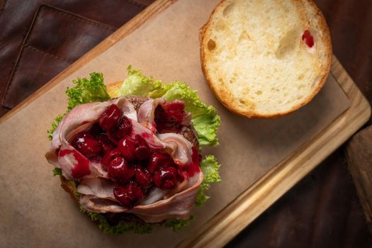 Fresh craft beef burger with fried slices of bacon and cherry sauce on top, fresh lettuce with lead bun laying next to it. Restaurant concept. Street food concept.