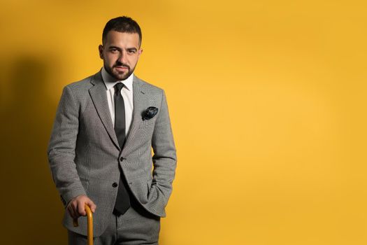 Old school trendy stylish handsome young man wearing grey suit, with one hand lifted standing with cane or umbrella in his hand isolated on yellow background.