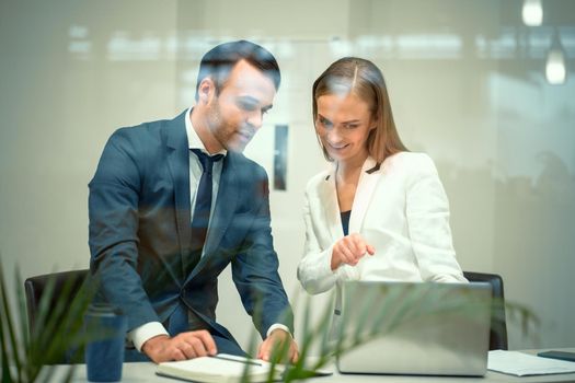Handsome young businessman and a woman working on laptop in the office. Smiling diverse business partners young man and woman discussing project sitting in a modern bright meeting.