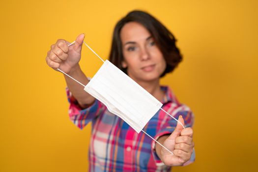 Cute young woman holds protective mask in foreground. Isolated on yellow background. Quarantine concept. Copy space.