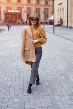 Outdoor portrait of charming spring time young french woman wearing sunglasses walking on camera with old town on background. Stylish female tourist standing at the city center square. Toned image.