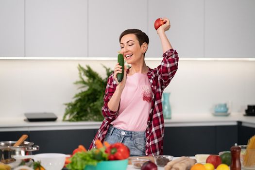 Young pretty housewife with a bob hairstyle cooking dinner or lunch standing in the kitchen choosing fresh vegetables smelling cucumber and apple in hand. Healthy food at home.