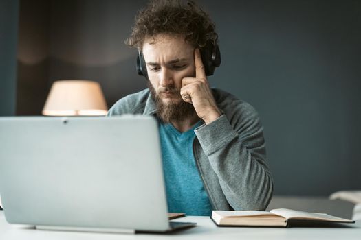 Serious young freelancer man working at laptop sitting at the desk at home office and taking notes. Remote working pensive young man working at home. Work from home concept.