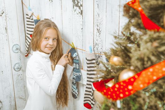 Cute Caucasian Girl in a White Jumper with Long Blond Hair Hangs Christmas Socks. Christmas Stockings. White Wood Background. Close-up. High quality photo