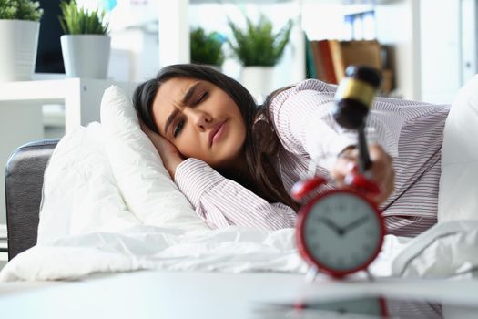 Portrait of young woman waking up in bad mood crash alarm with hammer. Female in pyjamas in bed at home, early start. Good morning, awake, angry concept