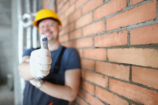 Close-up of foreman in helmet show thumbs up on construction site, handyman made good job. Worker standing near brick wall. Renovation, building concept