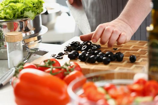 Close-up of man chef cutting olives on wooden cutting board, add ripe vegetable to salad. Person follow delicious new recipe. Cooking, hobby, taste concept